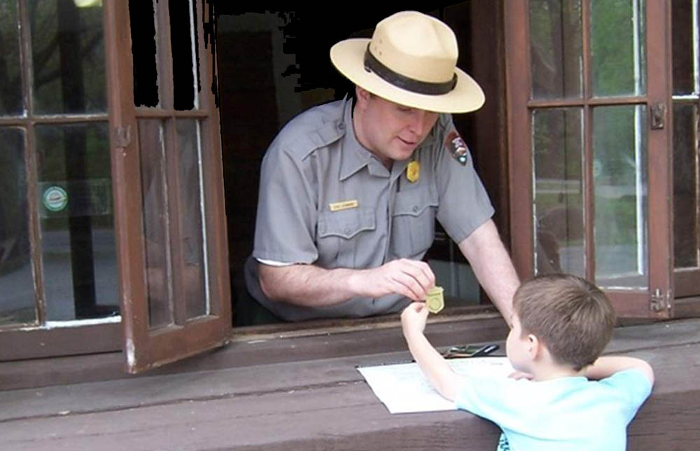 Man in broad brim hat and uniform with shield-shaped badge on his shirt leans out the Junior Ranger Station window to hand a boy a shield shaped badge.